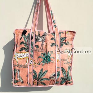 May include: A pink tote bag with a white and pink striped handle. The bag is made of quilted fabric and features a colorful print of animals and plants, including a zebra, a leopard, and palm trees. The text "Artist Couture" is printed on the bag.