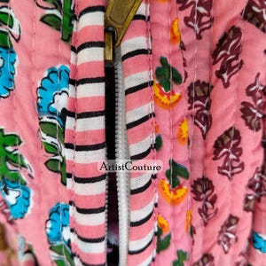 May include: Close-up of a pink quilted fabric with floral patterns and a brass zipper. The zipper is partially open, revealing a white and black striped fabric. The text "Artist Couture" is visible on the fabric.