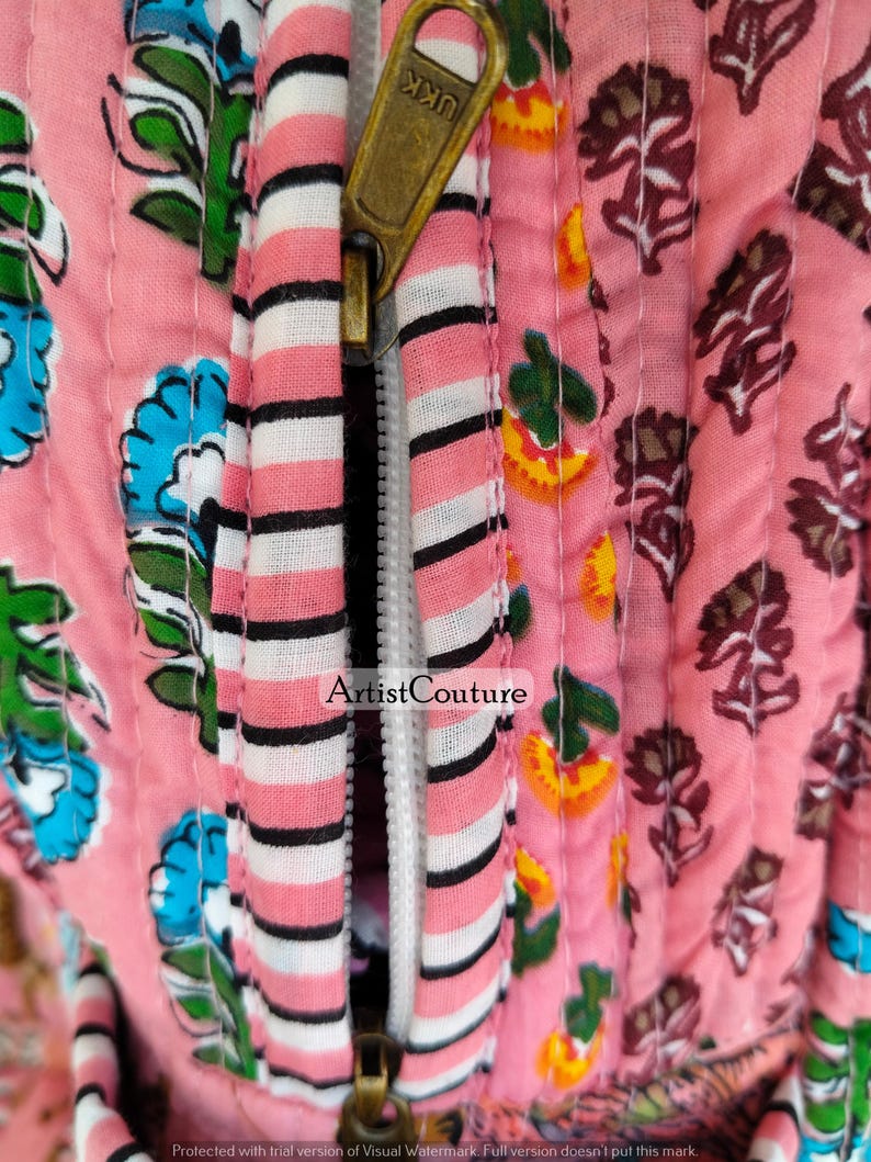 May include: Close-up of a pink quilted fabric with floral patterns and a brass zipper. The zipper is partially open, revealing a white and black striped fabric. The text "Artist Couture" is visible on the fabric.