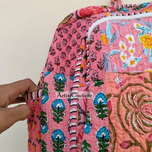 May include: Pink quilted backpack with floral and geometric patterns. The bag features a variety of designs, including blue floral motifs, and a large floral print. The bag has a white zipper and a handle at the top. The text "Artist Couture" is visible.