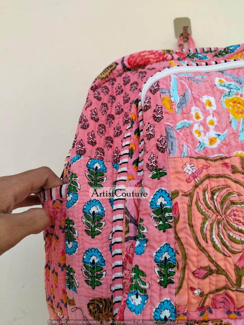 May include: Pink quilted backpack with floral and geometric patterns. The bag features a variety of designs, including blue floral motifs, and a large floral print. The bag has a white zipper and a handle at the top. The text "Artist Couture" is visible.