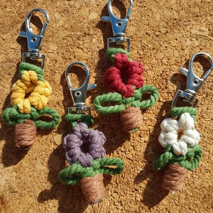 May include: Four handmade macrame flower keychains. Each keychain features a different colored flower: yellow, red, purple, and white. The flowers sit atop green leaves and brown pots, with silver-colored clasps for easy attachment.