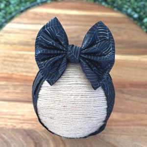 May include: A black baby headband with a large bow featuring a silver spiderweb design. The bow is centered on a beige fabric-wrapped base, perfect for Halloween or themed events. The headband is displayed on a wooden surface.