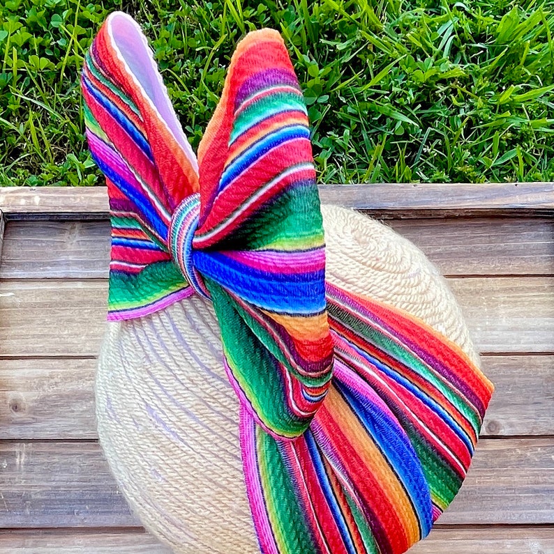May include: A large, colorful bow made from striped fabric in shades of red, green, blue, pink, and orange. The bow is tied around a beige, woven headband.