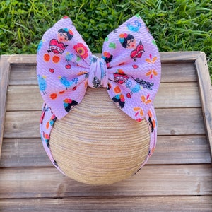 May include: A light purple baby headband with a large bow featuring a repeating pattern of Frida Kahlo portraits, hearts, flowers, and birds. The headband is wrapped around a tan, spherical form, set against a wooden background.
