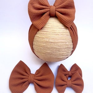 May include: Set of three brown fabric hair bows. The largest bow is attached to a brown headband. The bows are made of a textured fabric.