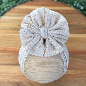 May include: A beige baby headband with a large, decorative bow. The bow features a delicate, eyelet lace design. The headband is wrapped around a beige, spherical form, suggesting it's for display or storage. The overall aesthetic is soft and feminine.