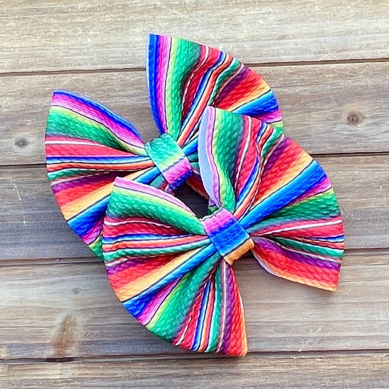 May include: Two colorful hair bows with a striped pattern reminiscent of a Mexican serape. The bows are made of a textured fabric and are tied in a classic bow shape.