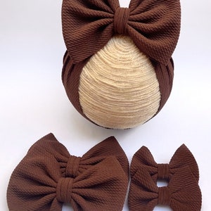 May include: Three brown fabric hair bows of different sizes. The largest bow is on a mannequin head. The bows are made of a textured fabric.