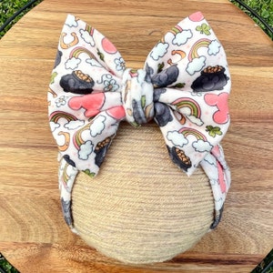 May include: A pink and white fabric bow with a rainbow, pot of gold, and shamrock print. The bow is tied in a knot and is ready to be worn in hair.