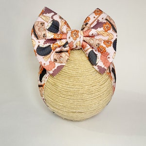 May include: A pink and orange headband with a bow featuring a Halloween-themed print of black cats, cauldrons, and stars.