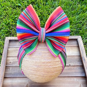 May include: A colorful striped bow headband with red, green, blue, and pink stripes. The headband is tied in a bow and is sitting on a wooden surface.
