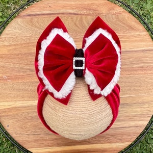 May include: Red velvet bow with white trim and a black buckle with rhinestones. The bow is attached to a brown headband.