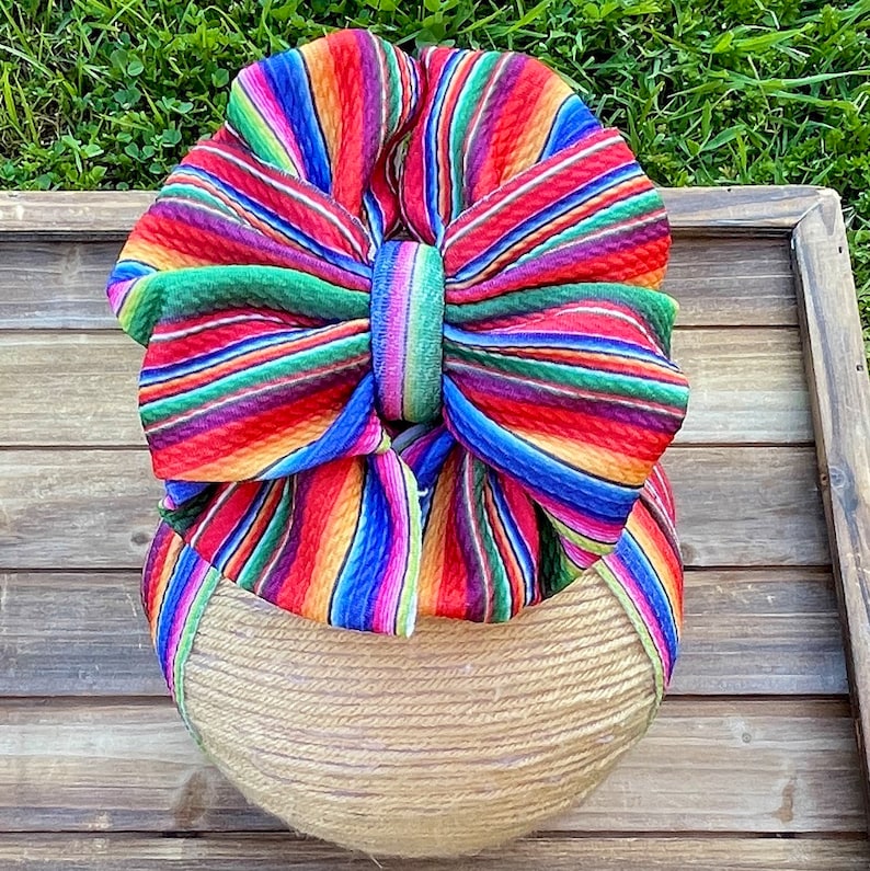 May include: A large, colorful bow made from fabric with a striped pattern in shades of red, orange, yellow, green, blue, and purple. The bow is tied around a round, woven base.