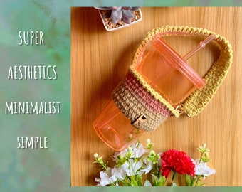 May include: A crocheted cup holder with a pink, beige, and yellow color scheme. The holder is attached to a long strap and is holding a clear plastic cup with a straw.