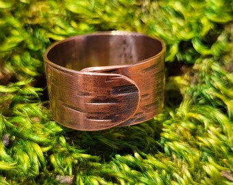 Puede incluir: Un anillo ancho de banda de cobre con textura martillada y borde redondeado. El anillo está sentado sobre un lecho de musgo verde.
