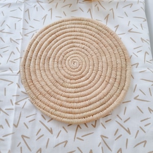 Op de afbeelding: Een ronde, geweven, naturelkleurige strooien placemat met een spiraalvormig ontwerp. De placemat ligt op een wit tafelkleed met een bruin abstract patroon.