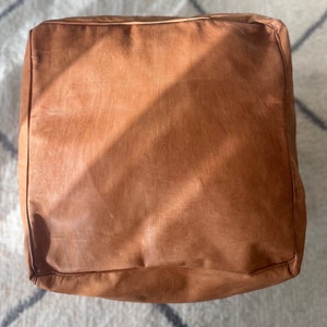 Puede incluir: Un puf otomano de cuero marrón de forma cuadrada. El puf está sobre una alfombra de color blanco y gris con patrones.