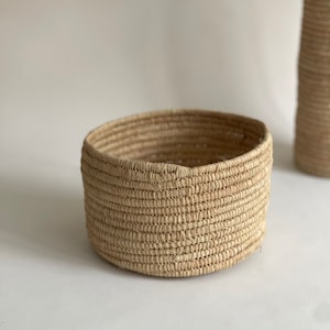 Op de afbeelding: Een ronde, geweven mand gemaakt van natuurlijke vezels. De mand is lichtbruin van kleur en heeft een gladde, gelijkmatige textuur.