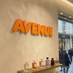 Puede incluir: La palabra "AVENUE" en letras naranjas está montada en una pared beige. Debajo, una estantería exhibe varios frascos de perfume. A través del cristal, se ve una escena de la calle con peatones y escaparates.