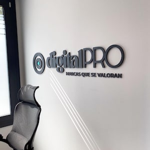 Könnte beinhalten: Ein schwarzes, dreidimensionales Wandschild mit dem Schriftzug digitalPRO, einem kreisförmigen Logo-Symbol auf der linken Seite und dem darunter stehenden Satz MARCAS QUE SE VALORAN. Das Schild ist an einer weißen Wand über einer Steckdose.