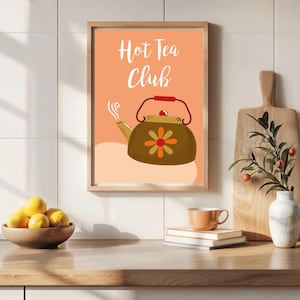 Può includere: Una stampa incorniciata con la scritta "Hot Tea Club" sopra un'illustrazione di una teiera marrone con manico rosso e motivo floreale. La stampa è su sfondo pesca. Una ciotola di legno con limoni, una tazza e un vaso con vegetazione sono su una superficie di legno.