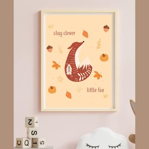 Può includere: Una stampa artistica incorniciata con un'illustrazione stilizzata di una volpe nei toni del marrone, bianco e rosso. L'opera include foglie autunnali, zucche e ghiande, con il testo "stay clever little fox". La stampa ha una cornice in legno chiaro.