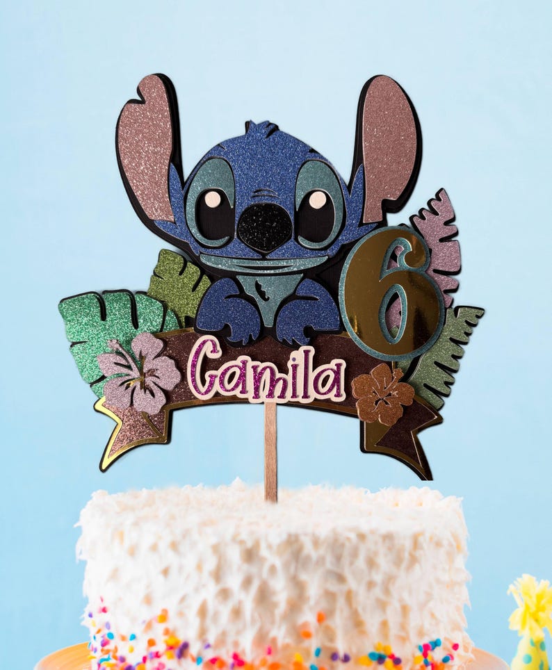 May include: A cake topper featuring a blue cartoon character with large ears, a black nose, and large eyes. The topper includes the number 6 and the name "Camila" on a banner, with tropical leaves and flowers. The topper is on a white cake with colorful sprinkles.