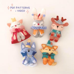 Può includere: Una collezione di bambole di animali in feltro fatte a mano, tra cui un gatto, un coniglio, un cervo, una volpe e una creatura blu simile a una volpe. Ogni bambola è adornata con costumi e accessori unici, come corone, mantelli e cappelli. Il testo "PDF PATTERNS + VIDEO" è visibile.