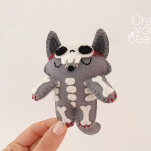 May include: Gray felt plush toy cat with a white skeleton design and red stitching. The cat has a skull face with black eyes and a small black nose. The toy is being held by a hand.
