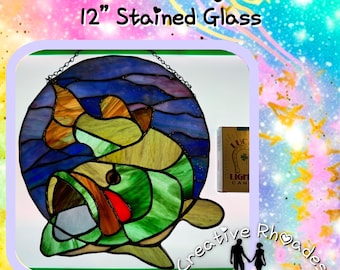 May include: A 12 inch round stained glass window depicting a bass fish in shades of green, blue, and orange. The fish is swimming in a blue and purple water scene. The window is hanging from a chain.
