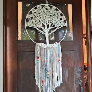 May include: A handcrafted macrame wall hanging featuring a tree design within a circular frame. The tree has green leaves and a white trunk. Below, strands of yarn in various colors, including blue, beige, and white, hang down, adorned with wooden beads.