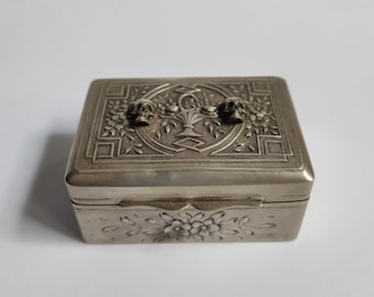 May include: A silver-coloured rectangular box with detailed floral and geometric patterns. The lid has a central floral design and two skull-shaped handles. The front of the box is decorated with a floral pattern. Likely used for jewellery or small keepsakes.