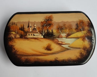 May include: A wooden box with a painted scene of a church and houses in a rural landscape. The church has a white steeple and a pink roof. The houses are white with brown roofs. The landscape is painted in shades of brown, yellow, and green. The box is signed "C.  Ostrovskaya" and "Konoval" in the lower corners.