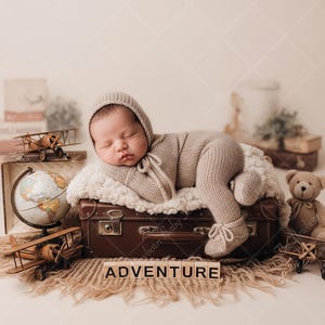 Puede incluir: Un bebé recién nacido durmiendo, con un conjunto y gorro de punto beige, sobre una maleta marrón. La escena incluye un globo terráqueo, aviones de juguete, un oso de peluche y la palabra "ADVENTURE" en bloques de madera, con temática de viaje.