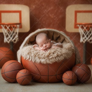 Puede incluir: Un bebé dormido descansa en un accesorio con forma de baloncesto, rodeado de otros balones. El fondo presenta aros de baloncesto y un fondo marrón texturizado, creando un escenario de fotos de recién nacidos con temática deportiva.
