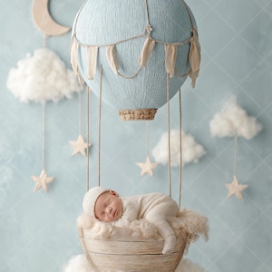 Könnte beinhalten: Ein schlafendes Baby in einem geflochtenen Korb, der an einem hellblauen Heißluftballon hängt. Der Ballon hat beige Akzente und hängt vor einem hellblauen Hintergrund mit Wolken, Sternen und einem Mond.