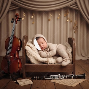 Könnte beinhalten: Ein Neugeborenes in einem beigefarbenen Strickoutfit und -mütze schläft in einem kleinen Holzbett. Eine Cello steht links, und eine Klarinette liegt unten. Noten und ein Notenblatt sind ebenfalls vorhanden.