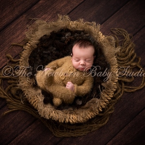 Könnte beinhalten: Ein Neugeborenes schläft in einem braunen geflochtenen Korb mit einem braunen Strick-Outfit. Der Korb ist mit braunem und schwarzem Material gefüllt. Das Baby trägt ein braunes Strick-Outfit mit einem Knopf vorne.