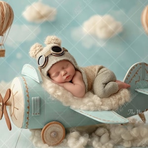 Puede incluir: Un recién nacido dormido con un gorro de aviador y un mono beige, descansando sobre un avión azul claro con una base de nubes blancas y esponjosas. Dos globos aerostáticos flotan en el fondo sobre un cielo azul.
