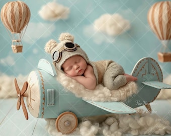 May include: A sleeping newborn in a beige aviator hat and jumpsuit, resting on a light blue airplane prop with a fluffy white cloud base. Two hot air balloons float in the background against a sky blue backdrop.