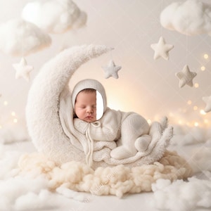 May include: A newborn baby in a white outfit and cap is nestled on a fluffy, white crescent moon prop. The scene is decorated with soft clouds and hanging stars, creating a dreamy, celestial theme.