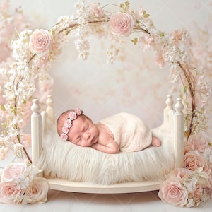 May include: A newborn baby sleeps peacefully on a small, white bed with a fluffy, white blanket. The bed is surrounded by pink flowers and a floral arch. The baby wears a pink floral headband and is wrapped in a soft, cream-colored blanket.