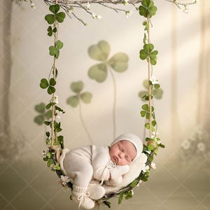 May include: A newborn baby wearing a white knit outfit and cap, sleeping peacefully in a decorative swing adorned with shamrocks and white flowers. The swing is suspended from a branch with more shamrocks and flowers.