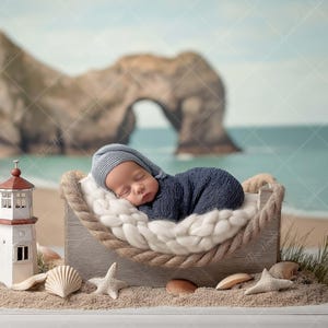 May include: A newborn baby wearing a blue outfit and hat, sleeping peacefully in a wooden cradle with a rope border. The cradle rests on sand with seashells and a miniature lighthouse. The background features a beach scene.