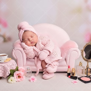 Pode incluir: Um bebé a dormir, vestido com um conjunto rosa de malha, incluindo um roupão, chapéu, luvas e botinhas, descansa num pequeno sofá rosa. Um serviço de chá em miniatura, flores e artigos de maquilhagem estão dispostos por perto.