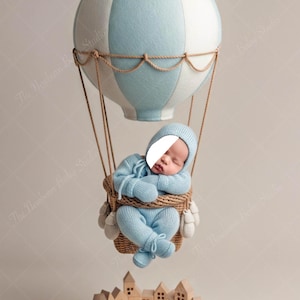 Puede incluir: Un bebé recién nacido con un conjunto y gorro de punto azul claro, durmiendo en una cesta de mimbre suspendida de un globo aerostático azul y blanco. Un pueblo de madera se encuentra debajo. The Newborn Baby Studio está escrito en la imagen.