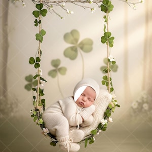 Può includere: Un neonato addormentato, vestito con un completo in maglia color crema e un cappello abbinato, riposa su un cuscino di maglia in un'altalena decorata con trifogli e fiori. L'altalena è sospesa a un ramo.