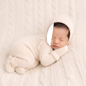 May include: A cream-coloured knitted newborn outfit consisting of a long-sleeved romper with footies and a matching bonnet. The romper features delicate tie closures at the side and the bonnet frames the face, both crafted in a soft, fine-gauge knit.
