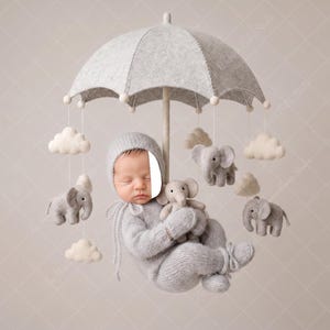 May include: A newborn baby in a gray knit outfit and hat, cradling a small elephant plush toy. The baby is suspended beneath a gray felt umbrella mobile with felt clouds and elephant ornaments. The background is a soft, neutral color.
